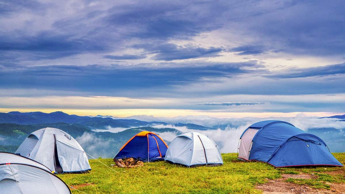Plusieurs tentes bleues et blanches dressées sur prairie verdoyante, chaîne montagneuse en arrière-plan sous ciel nuageux Plusieurs tentes bleues et blanches dressées sur prairie verdoyante, chaîne montagneuse en arrière-plan sous ciel nuageux